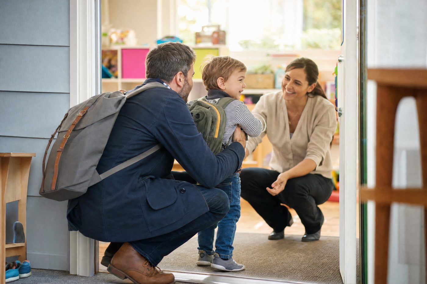 A dad crouching at a bright childcare center doorway, handing off his toddler with a small backpack to a smiling caregiver