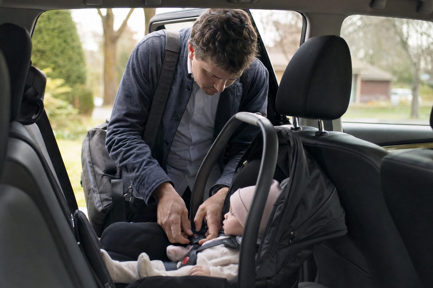 A dad with a work bag on his shoulder buckling his infant into a rear-facing car seat early on a weekday morning